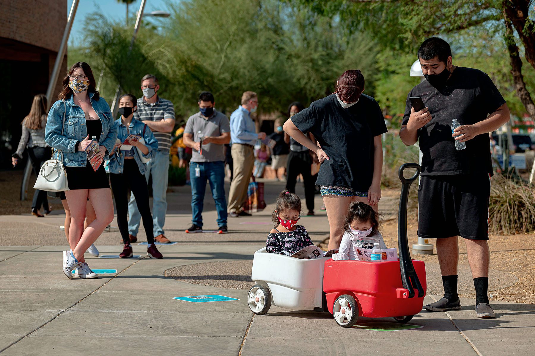 People wait in line outside a polling place including a family in the foreground with two little girls in a toy wagon.