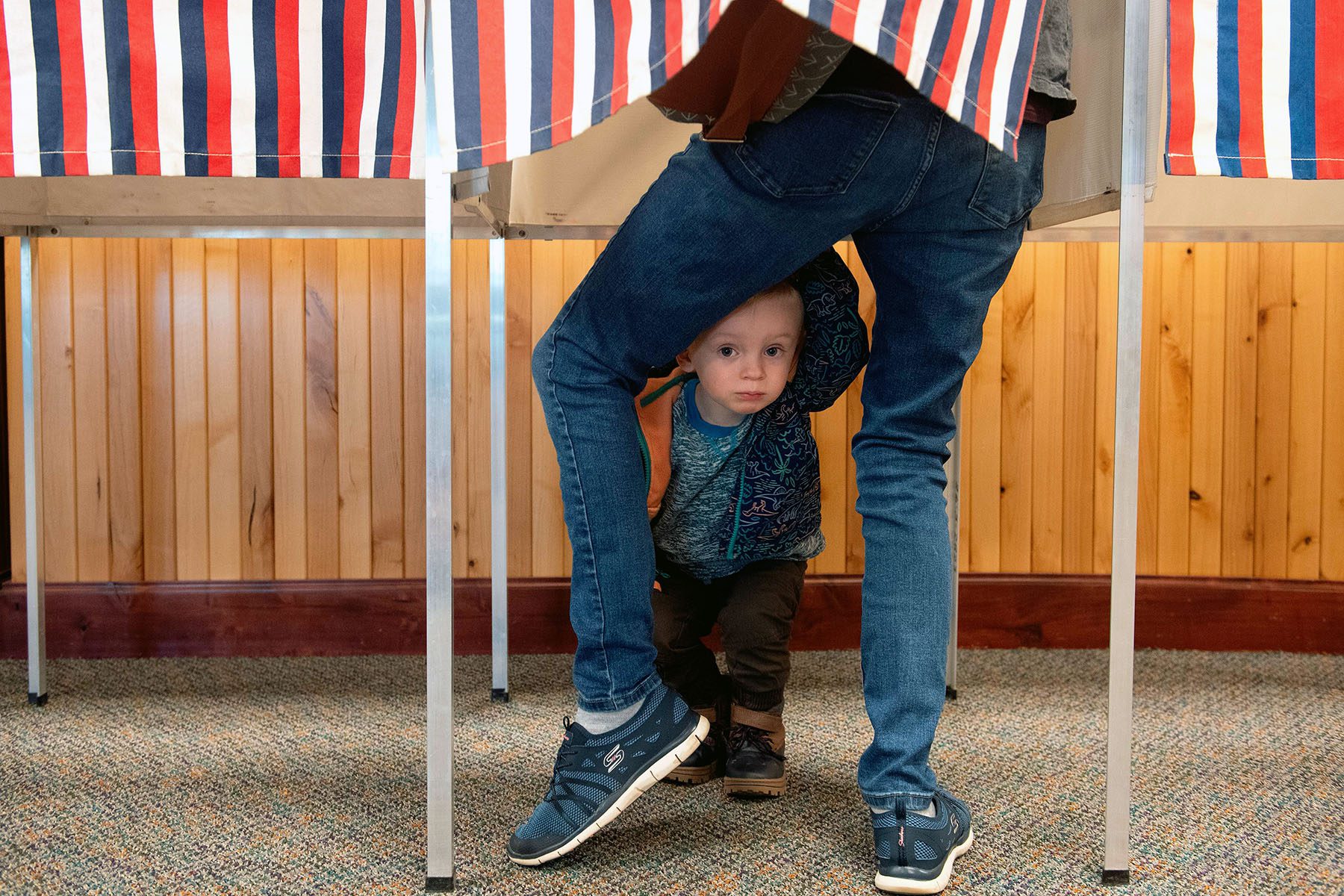 A child peaks at the photographer from under his mother's legs as he waits for his mother to cast her ballot.