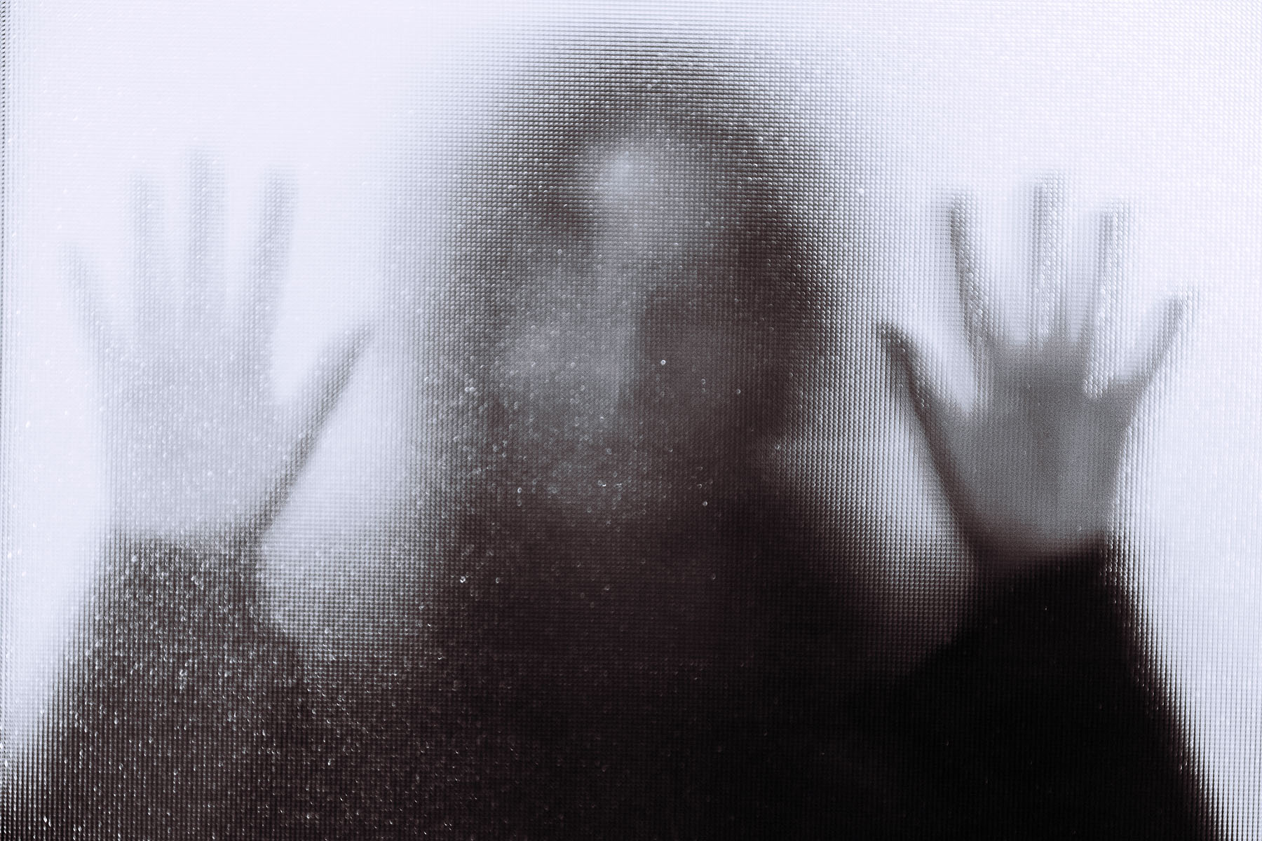 Back lit image of the distorted silhouette of a person with their hands pressed against a glass window.