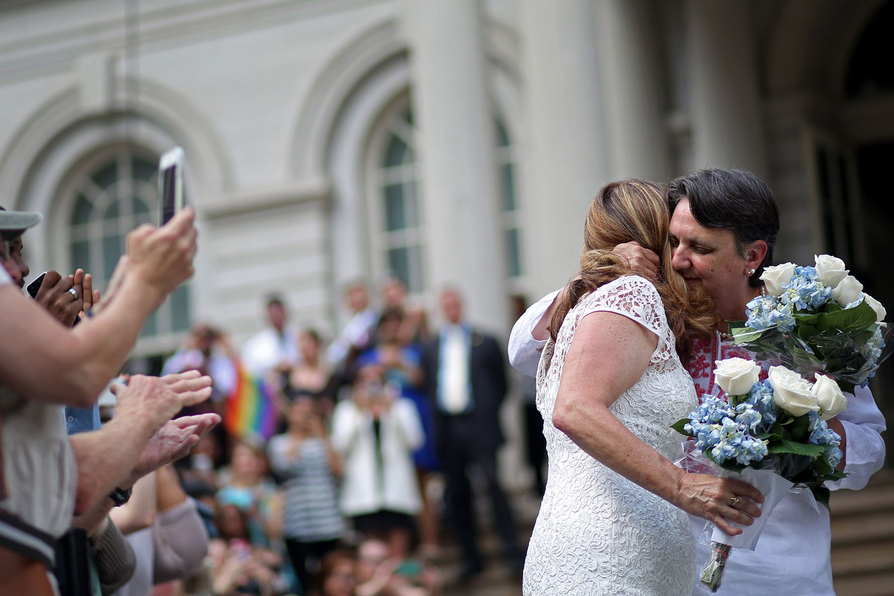 Two women embrace after getting married as people around them clap, cheer and take pictures.