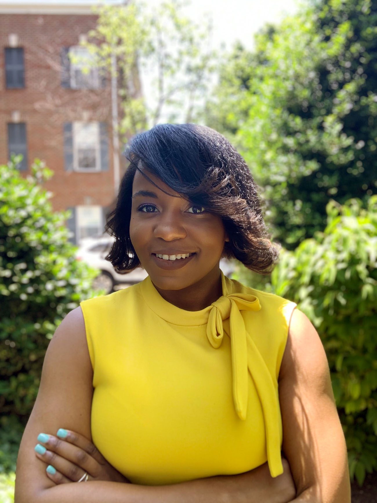 Smiling portrait of Kenyora Lenair Parham crossing her arms. She is wearing a yellow shirt with a bow and is surrounded by greenery.