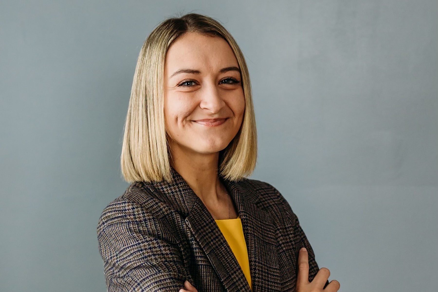 Anderson Clayton smiles as she poses for a portrait. She is wearing a checkered blazer and a yellow shirt.