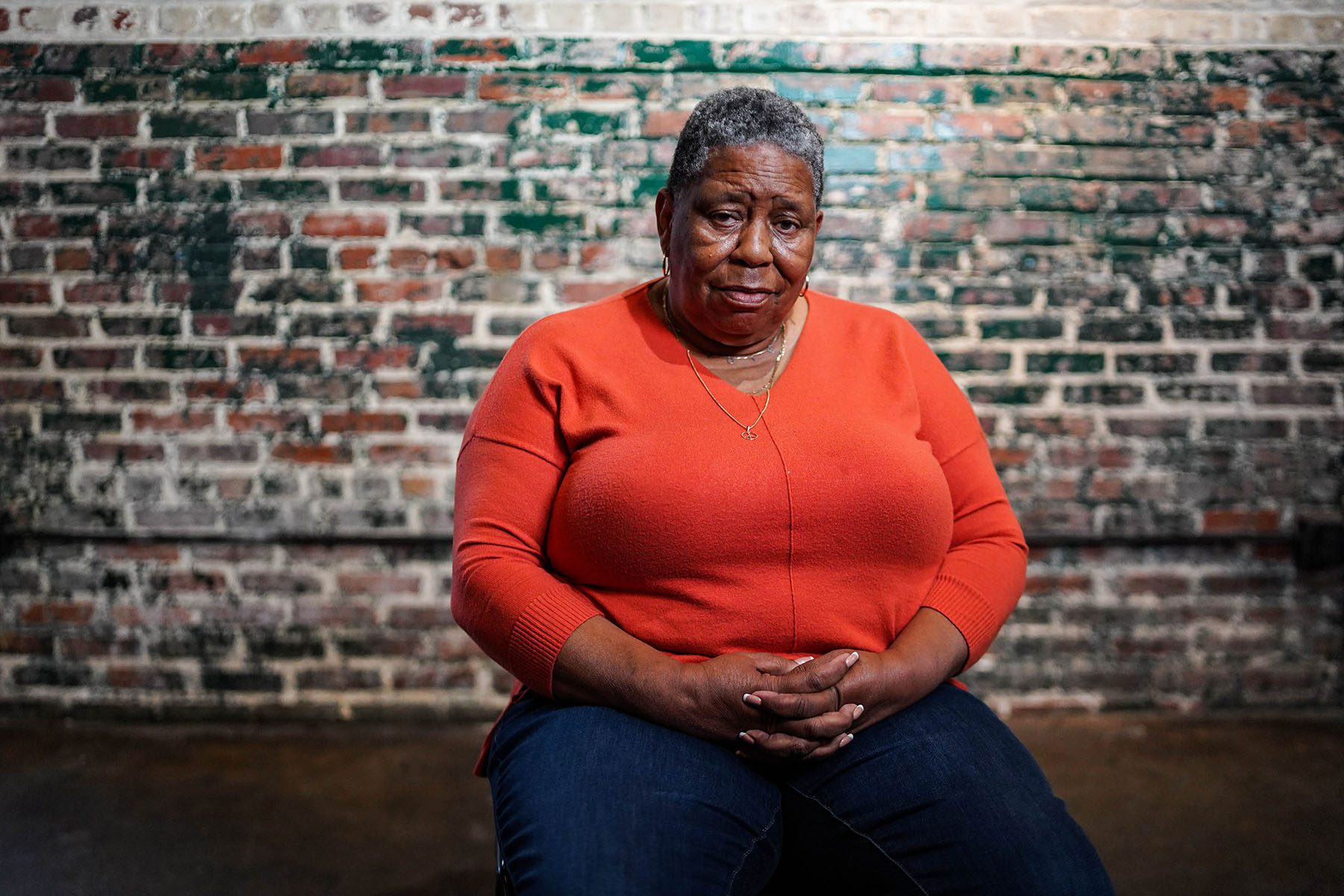 JoAnne Bland smiles slightly as she sits down and poses for a portrait. She is wearing an orange sweater and sitting in front of a brick wall.