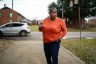 JoAnne Bland raises her hand as she speaks to somebody off screen. She is standing on a sidewalk in a neighborhood.