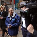 Chicago Mayor Lori Lightfoot walks on a city street with a man holding a camera in the foreground