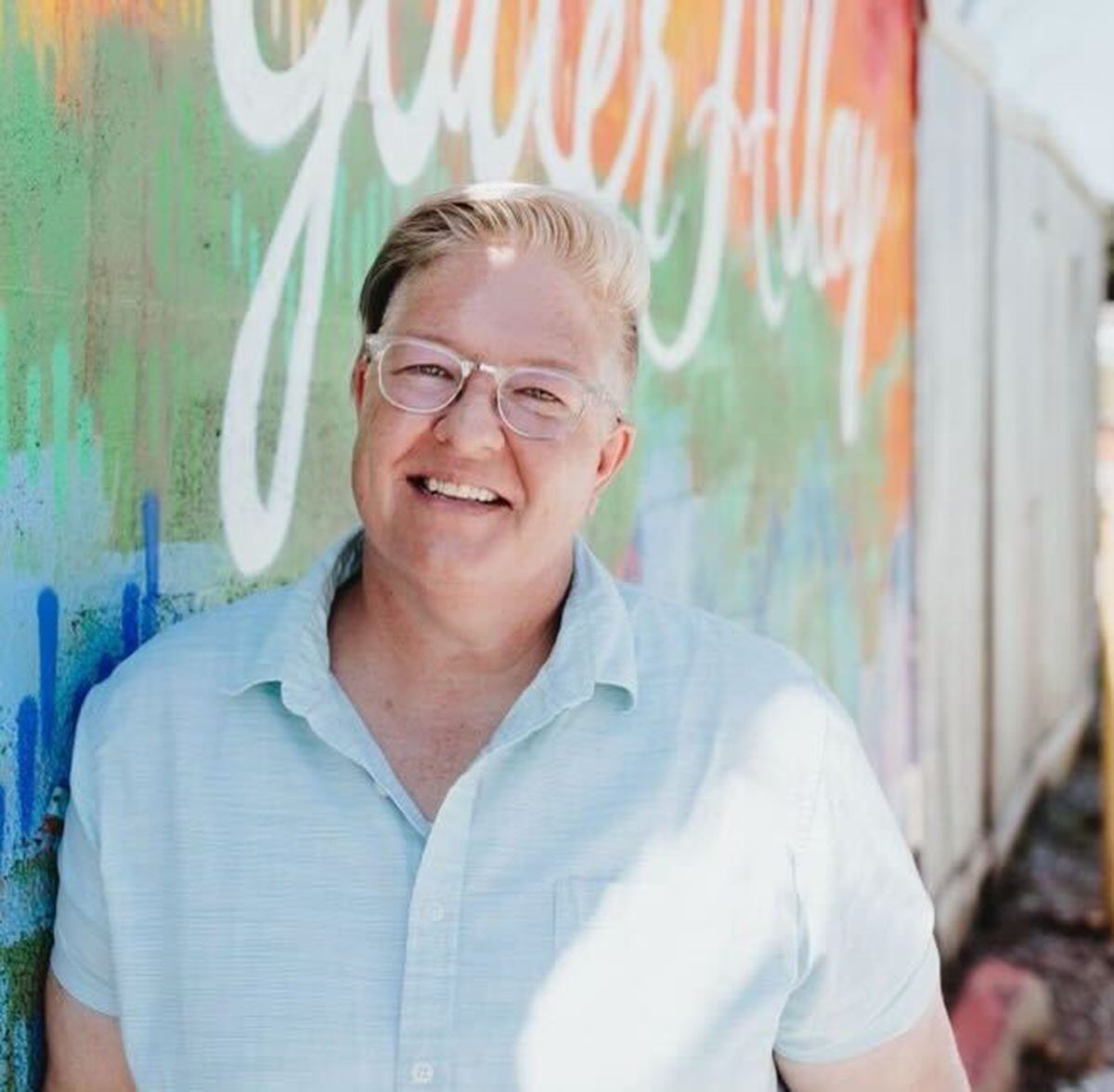 Portrait of Kris Williams posing against a colorful wall. She is wearing glasses and a light blue shirt.