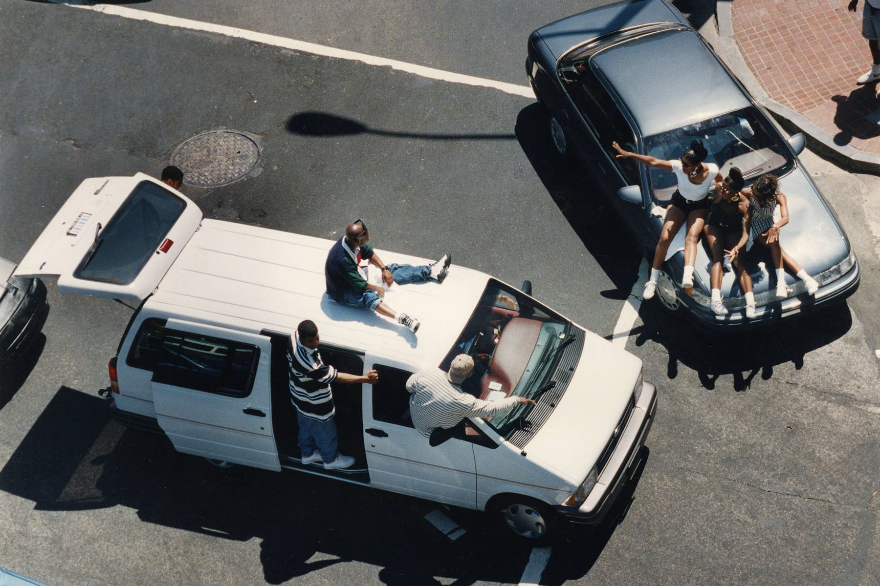 Two cars at a stop at an intersection with freaknik attendees sitting on top of cars.