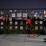 people walk by a fence at night time that has the photos of people killed by the police posted on it.