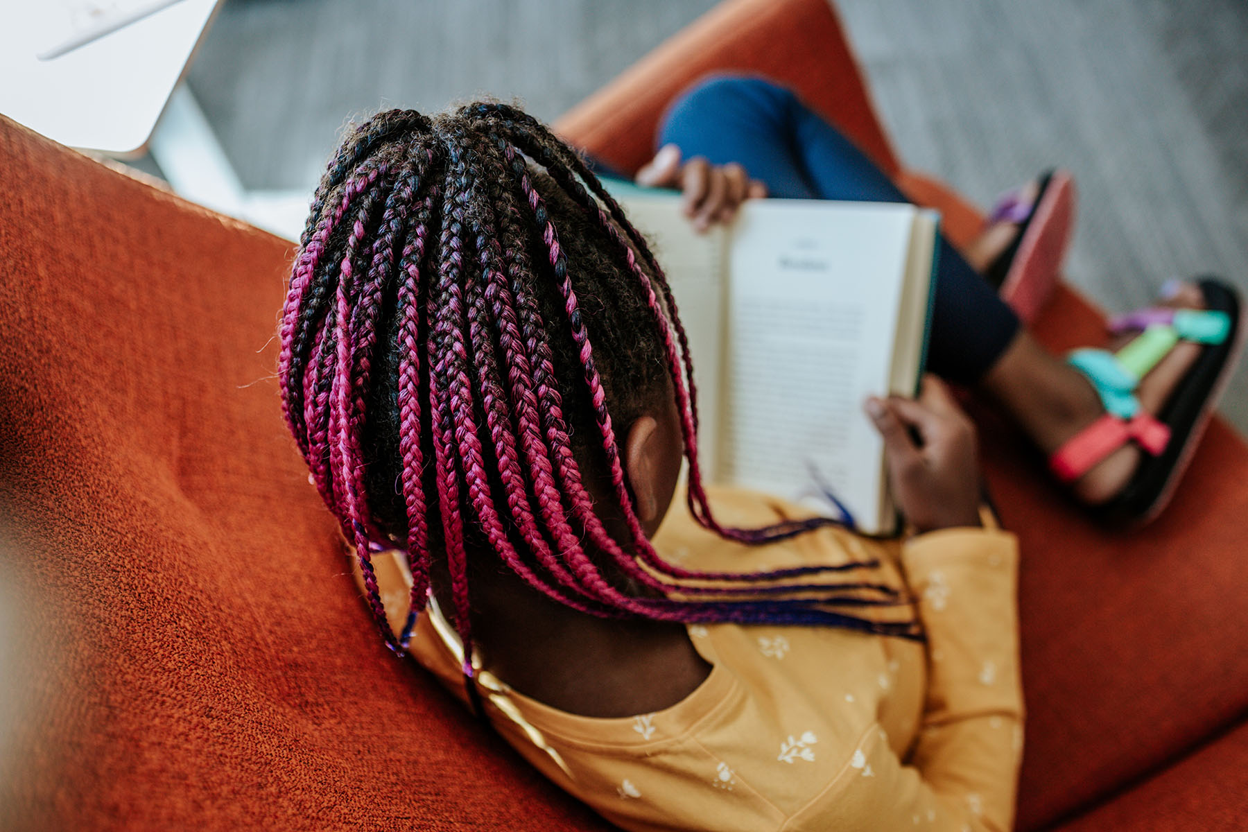 Young black girl reading a book on an orange chair.