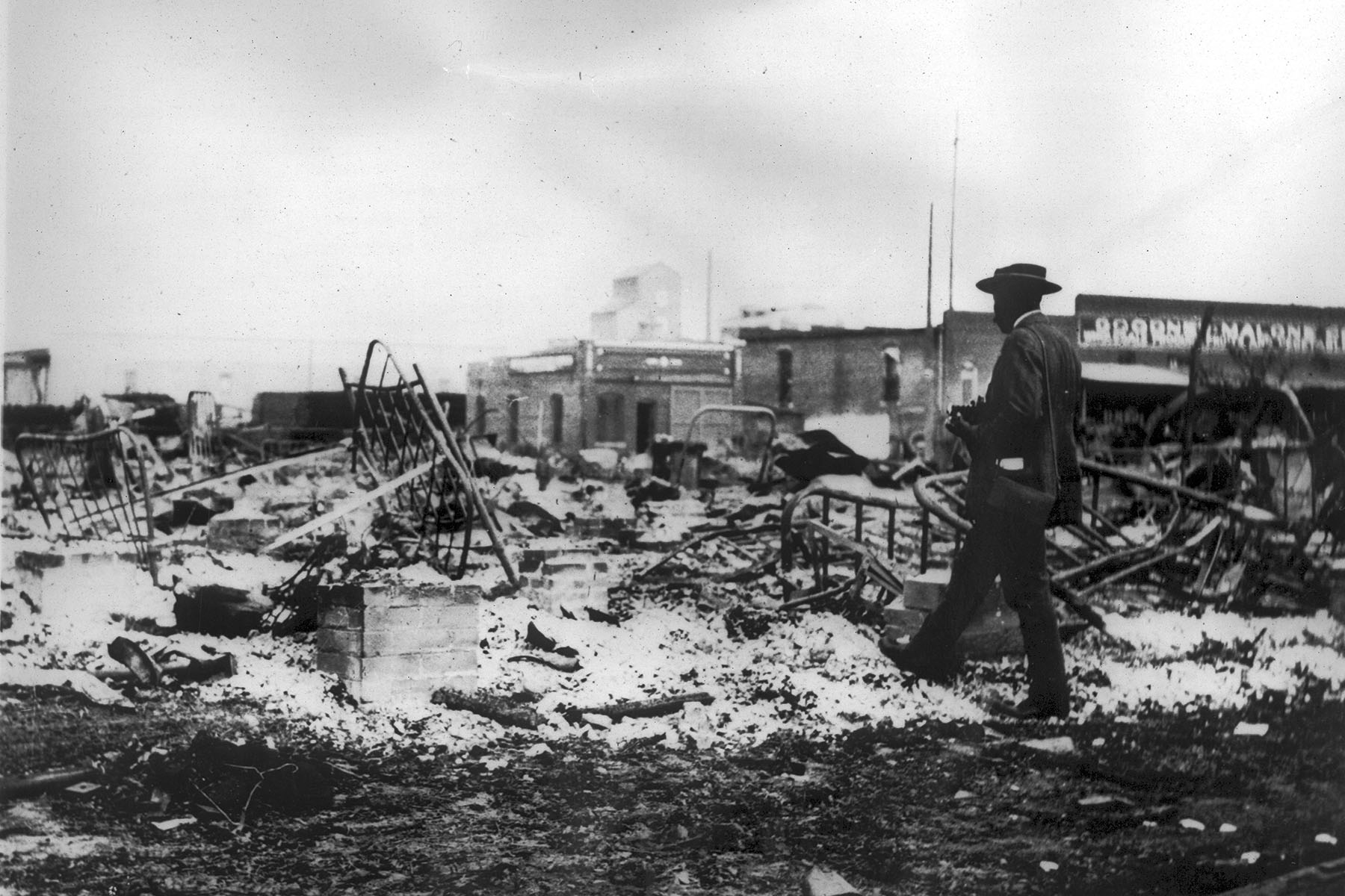 A black man with a camera looks at the skeletons of iron beds which rise above the ashes of a burned-out block after the Tulsa Massacre.