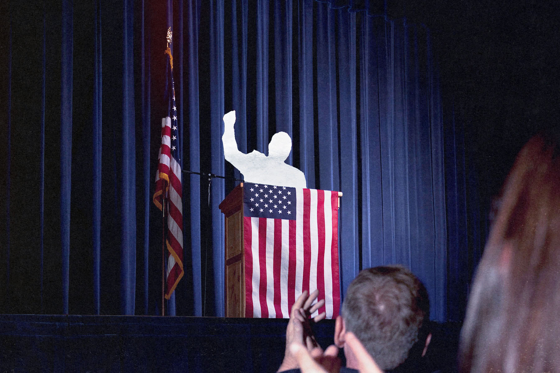 Photo collage of a silhouetted figured raising their fist while standing at a podium as people clap in the foreground. The figure at the podium is surrounded by American flags.