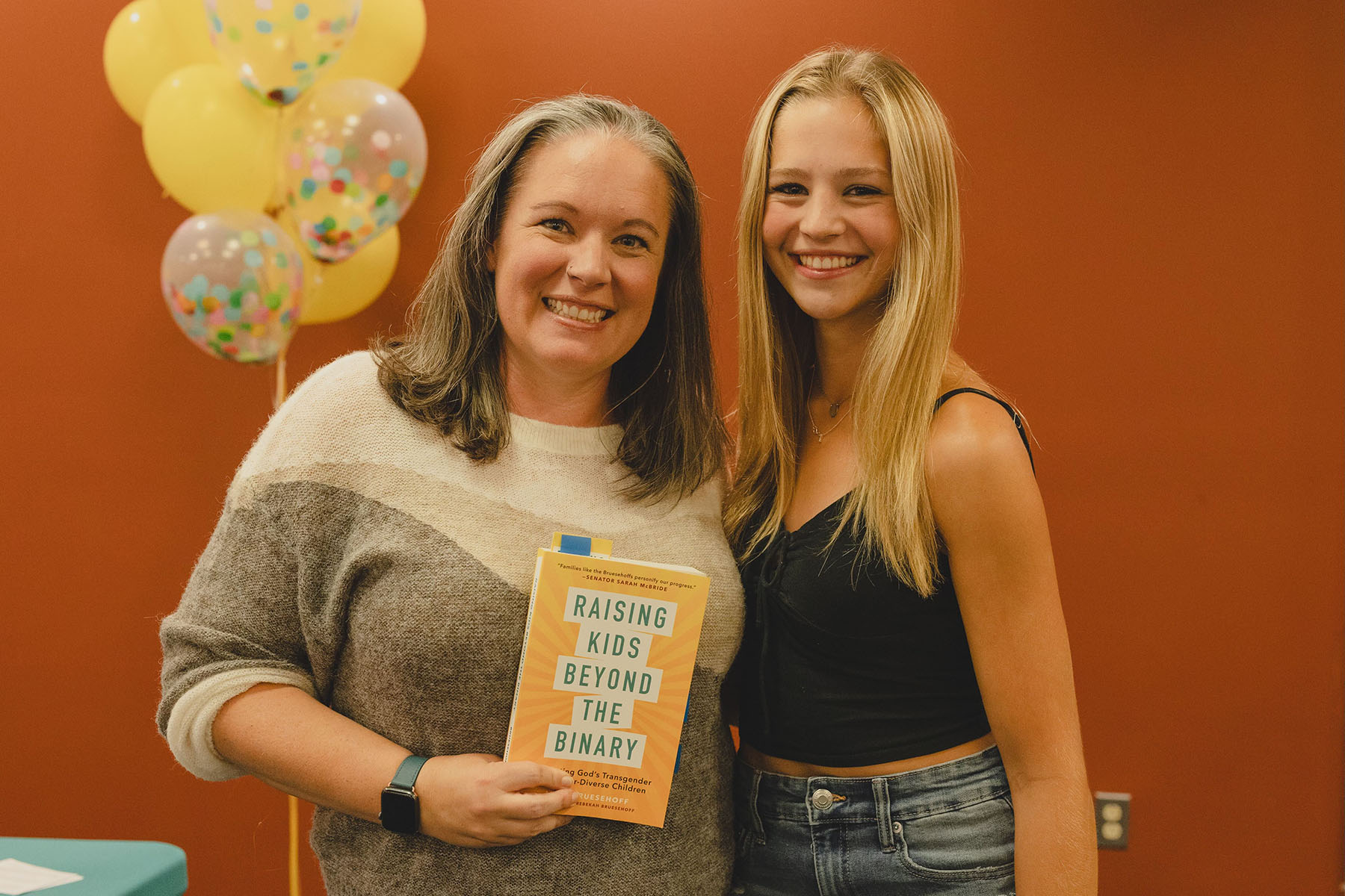 Jamie and her daughter Rebekah smile as they pose for a picture. Jamie is holding her book "Raising Kids Beyond The Binary."