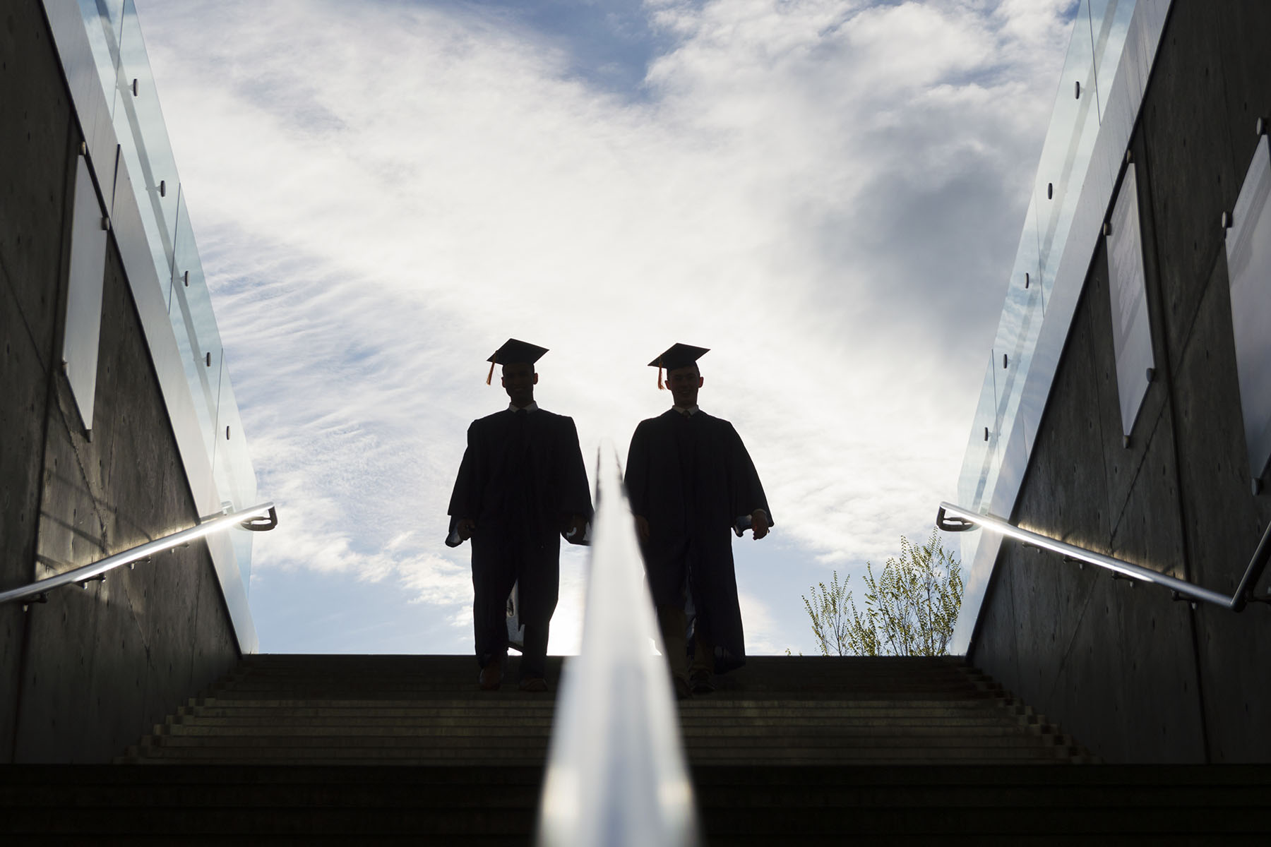 the silhouette of two college graduates in cap and gown climbing up a set of steps.