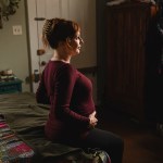 Sara Toups sits on a bed and holds her pregnant belly at her home.