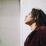 Non-binary person with eyes closed leaning on bathroom wall.