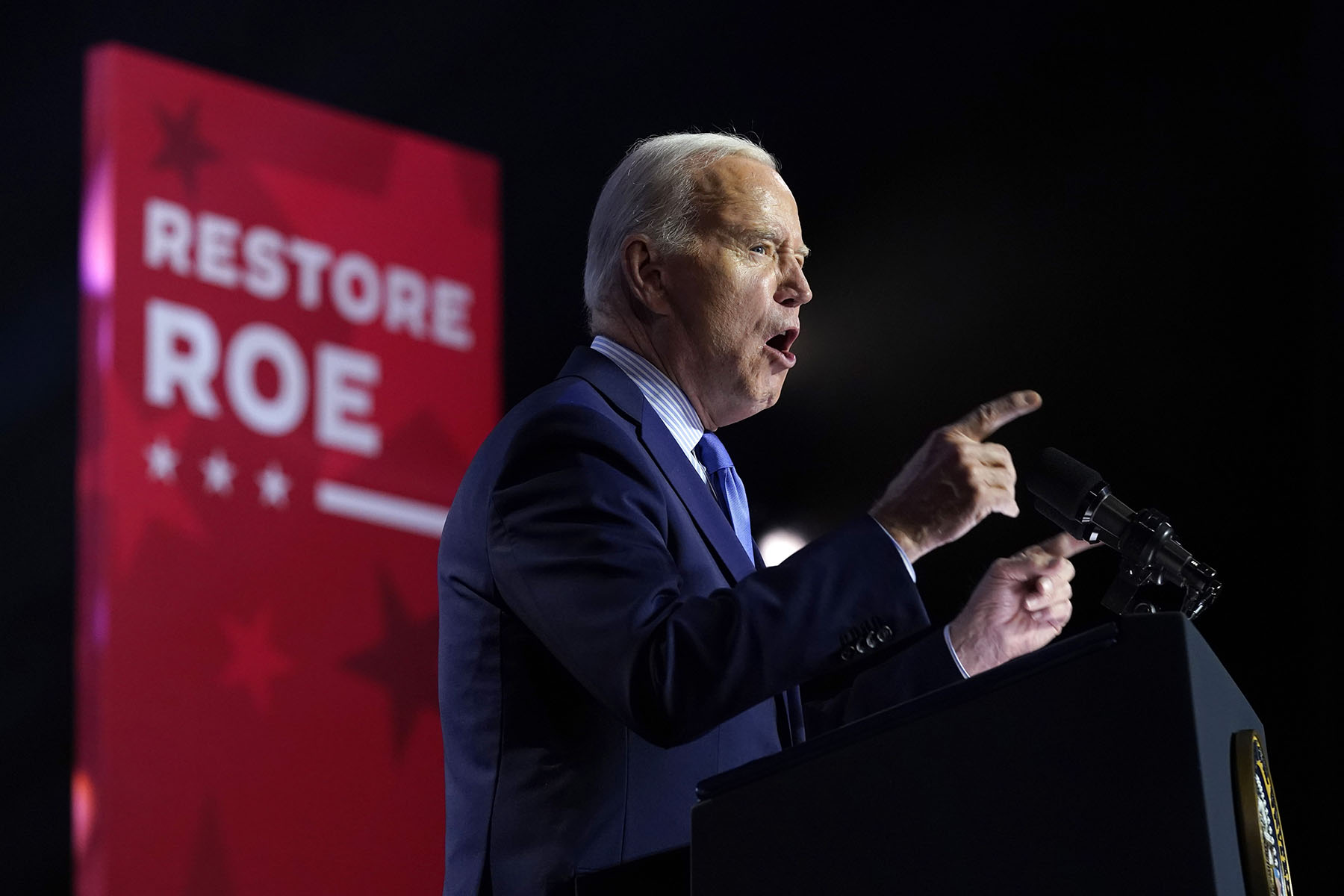 President Biden speaks during an event. Behind him a panel reads "Restore Roe."