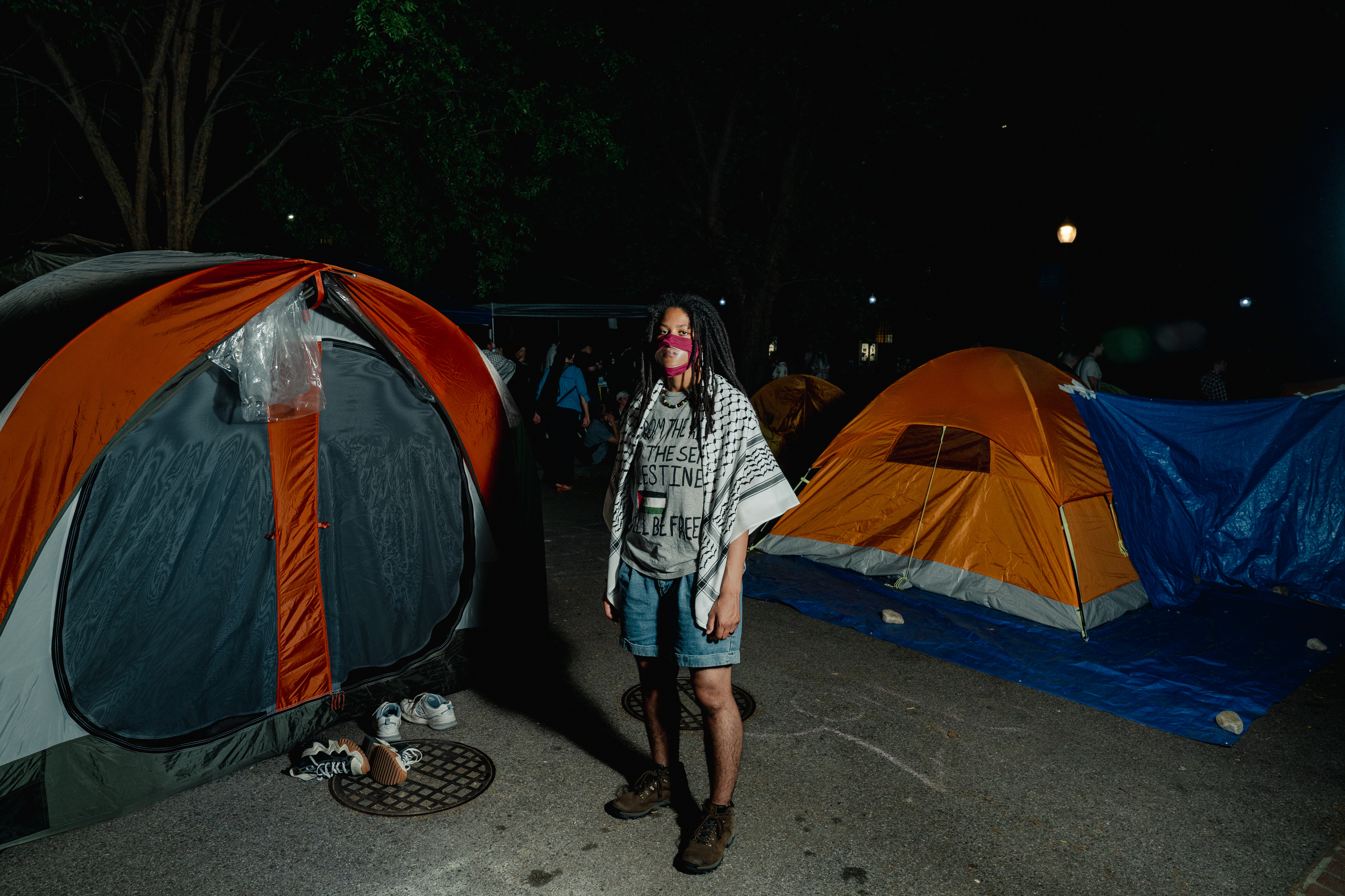 Bobbi-Angelica Morris poses for a portrait amidst tents at the George Washington University encampment. They are wearing a clear face mask, a keffiyeh is draped on their shoulders, and they are wearing a T-shirt with a Palestinian flag on it.