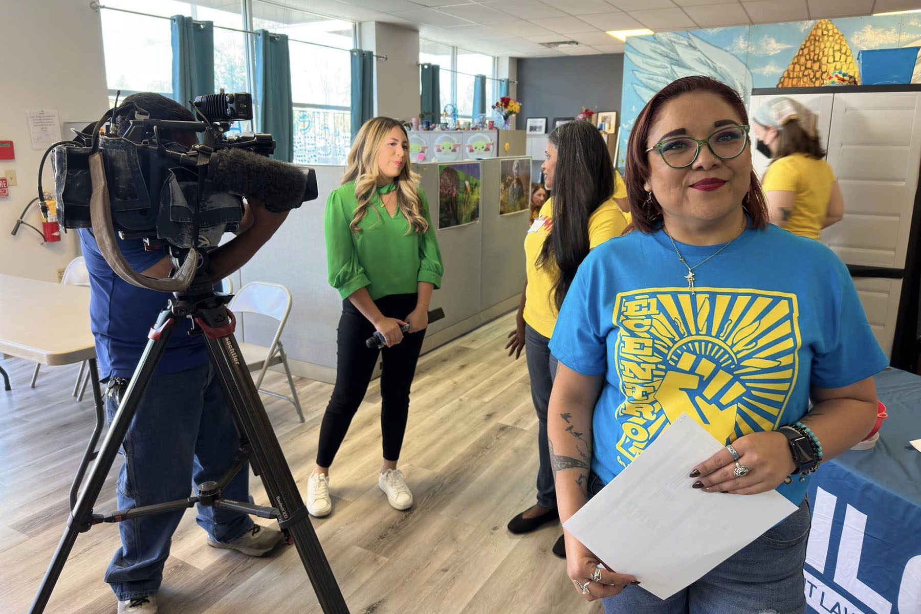 Fabiola Landeros smiles as a news camera films an interview behind her. She wears a blue shirt with a raised fist on it that reads "El Centro."