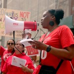 Union members are seen rallying. Many hold signs and one in the foreground chants into a megaphone.