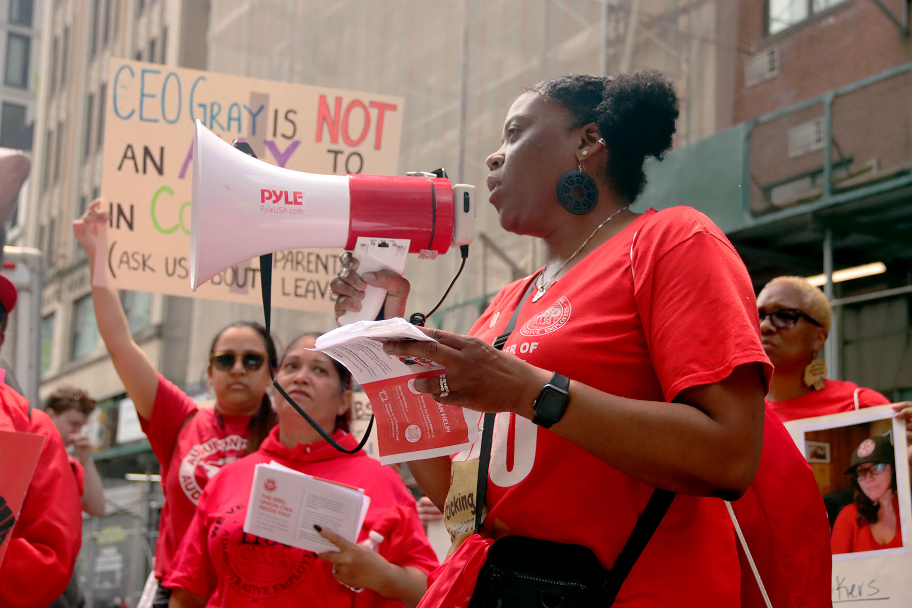 Union members are seen rallying. Many hold signs and one in the foreground chants into a megaphone.