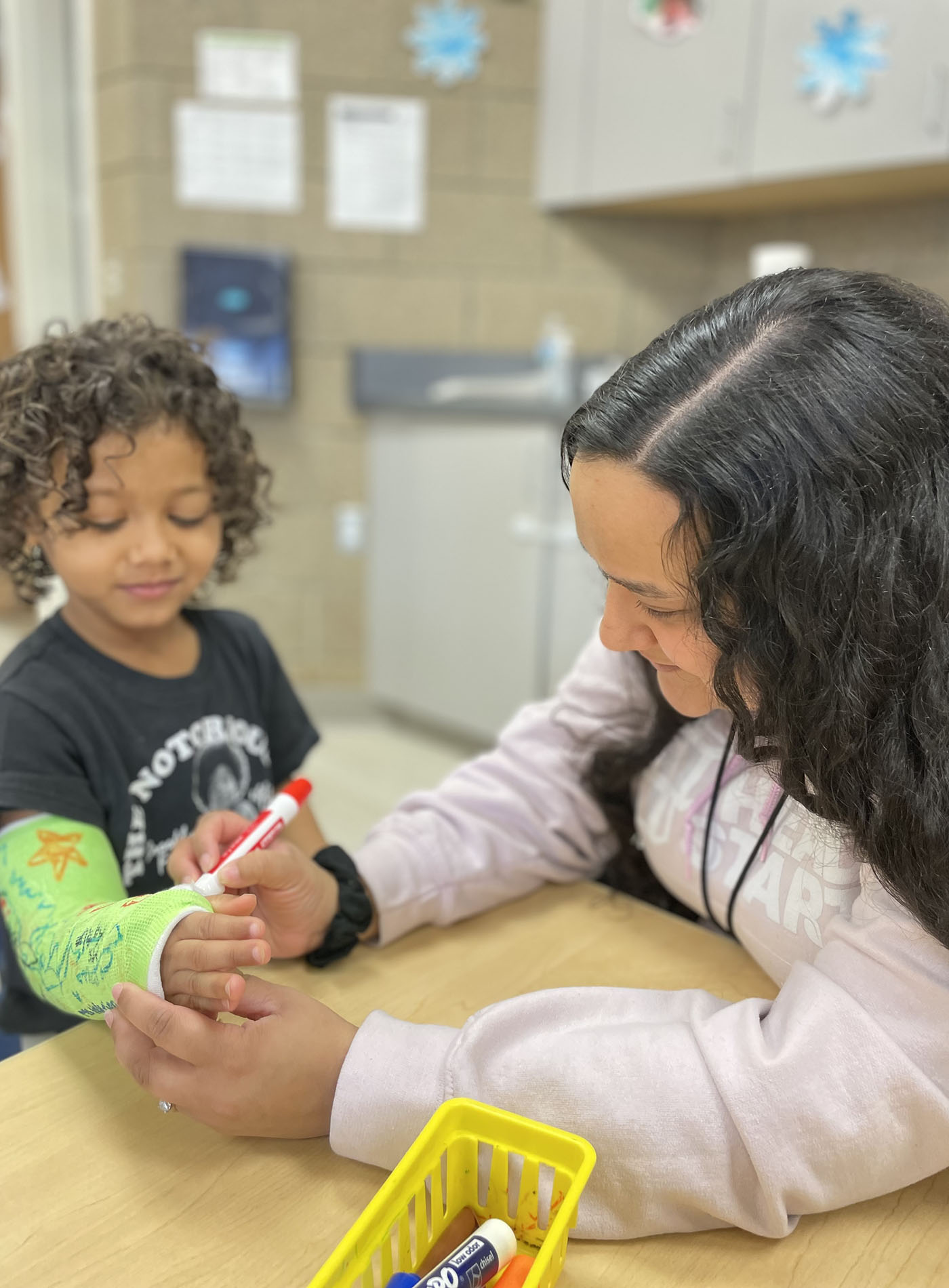 Katelyn Sarkar draws on a student's cast in a classroom as they both smile.