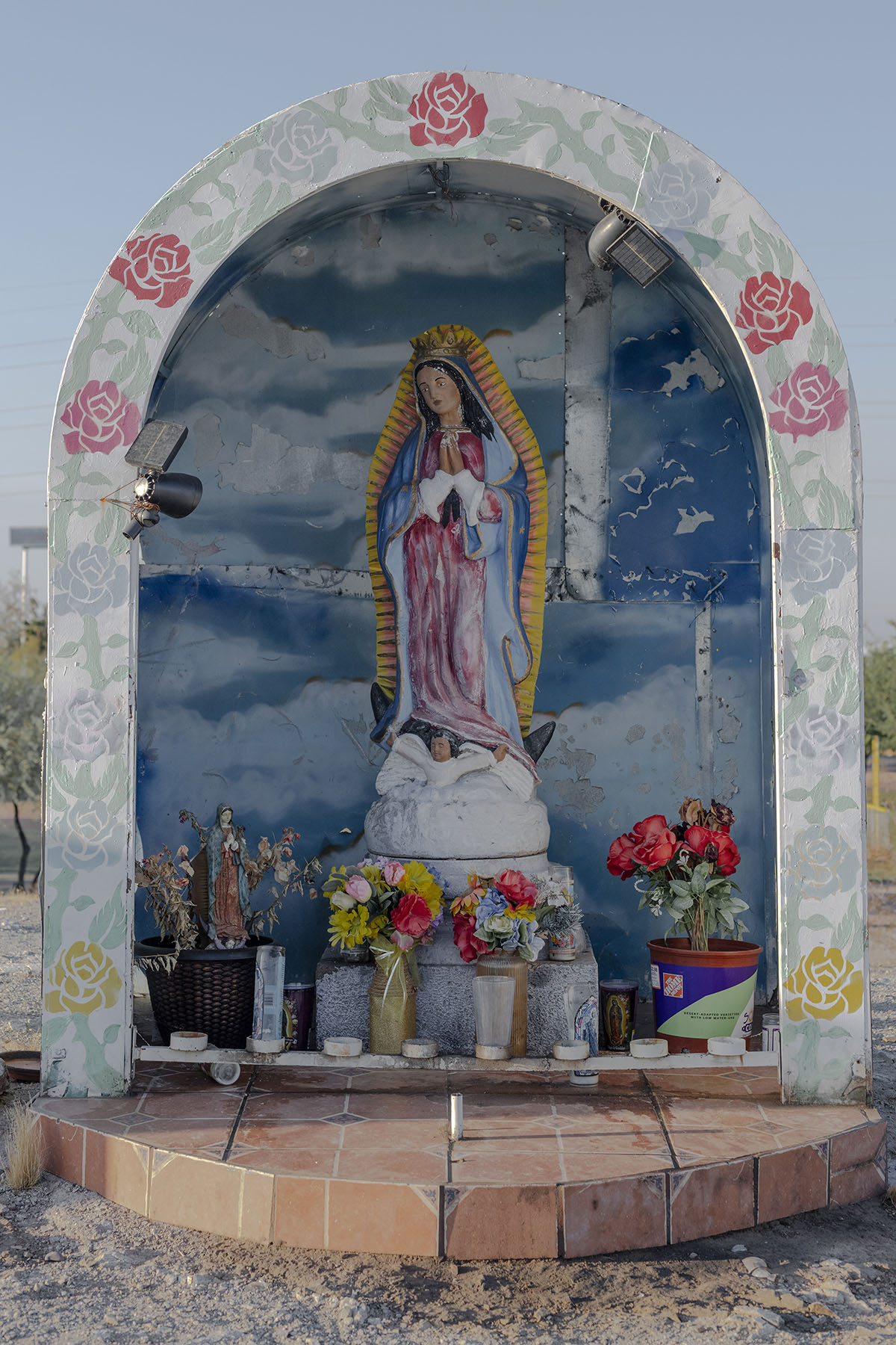A painted statue of Our Lady of Guadalupe is displayed in a decorative niche surrounded by flowers and candles, set against a blue sky background.