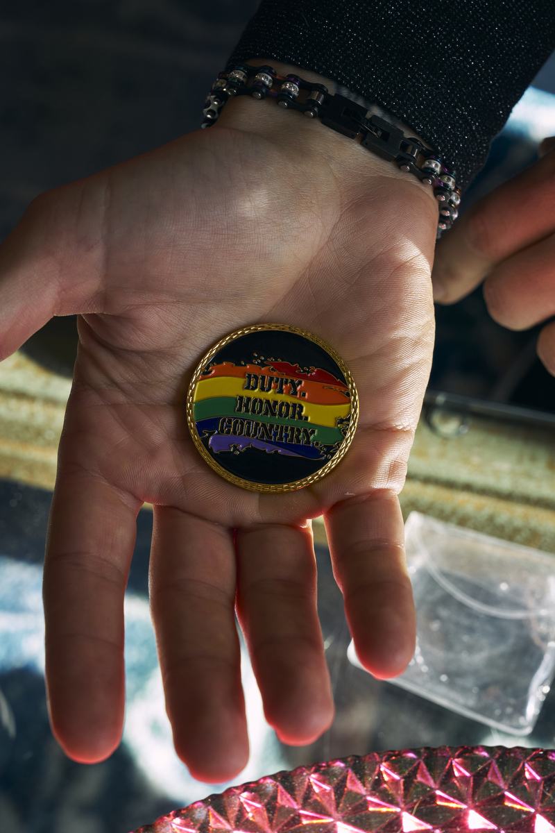 A hand holds a medallion that says duty, honor, country over a rainbow motif.