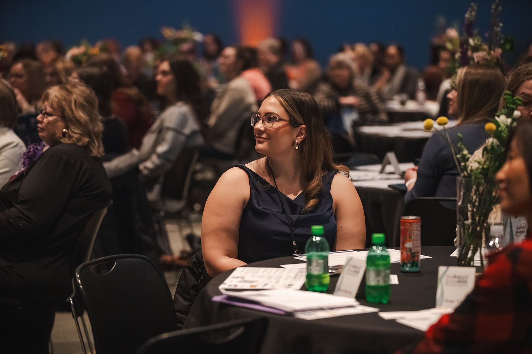 A woman seated at a table smiles as she listens to speakers during the We Rally & We Rise Women’s Conference.