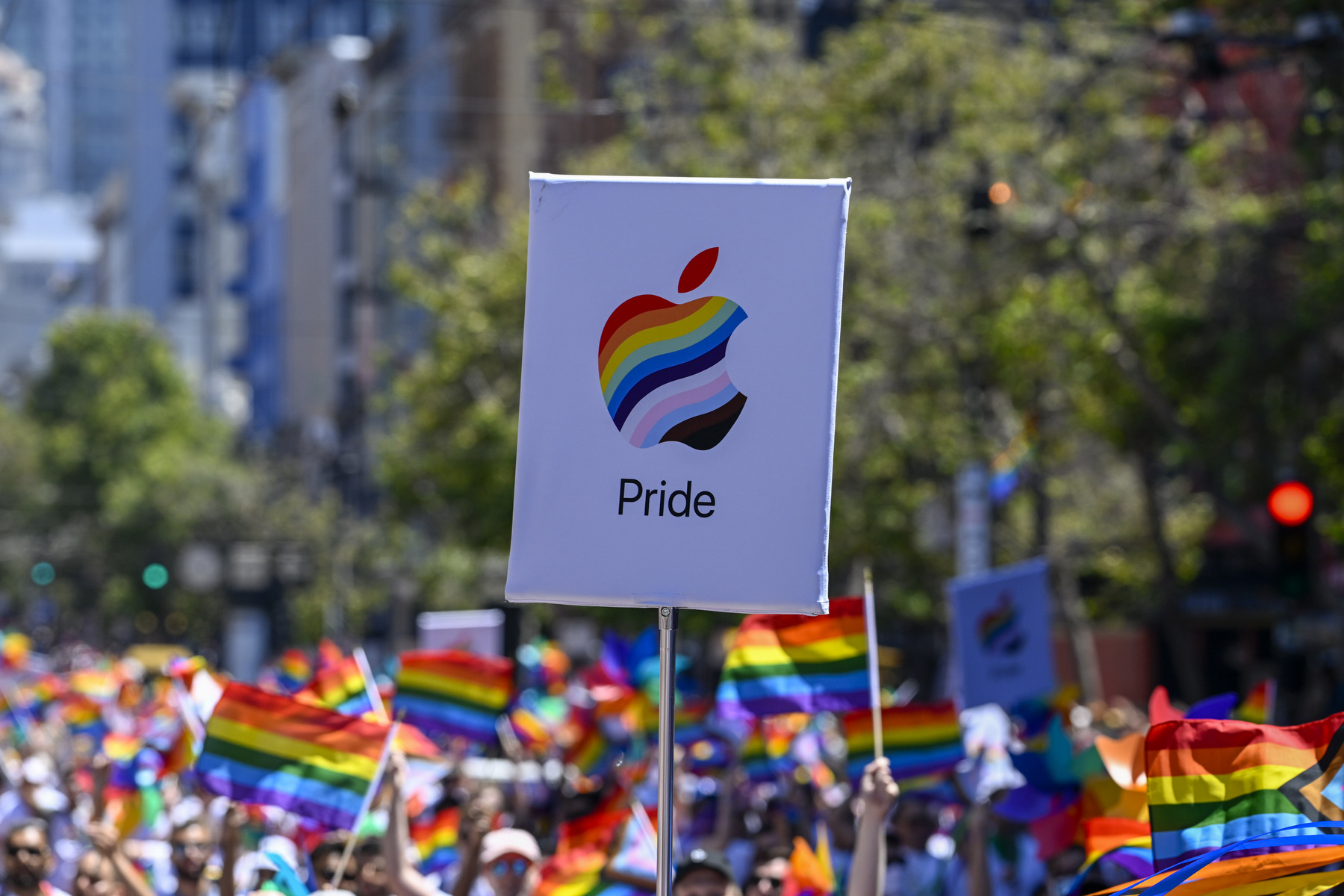 A sign with an Apple logo in the pride flag colors is raised above a crowd.