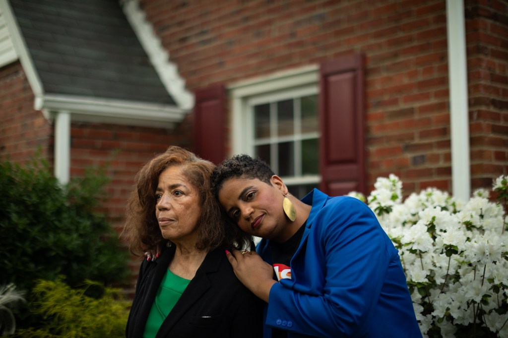 Two women standing together in front of a house. One woman is leaning on the other.
