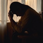 Silhouetted woman sits with her head in her hands in a dimly lit room.