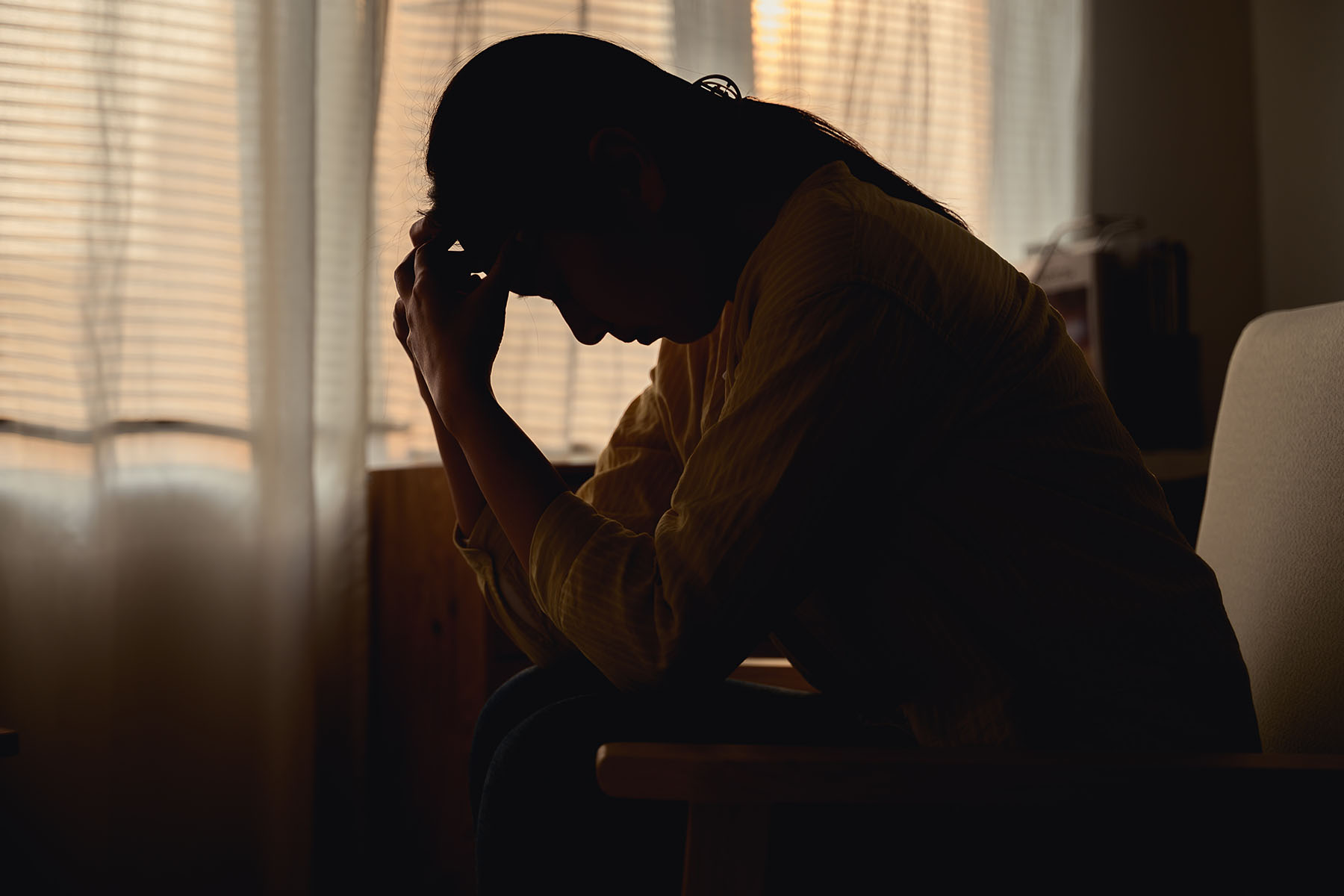 Silhouetted woman sits with her head in her hands in a dimly lit room.