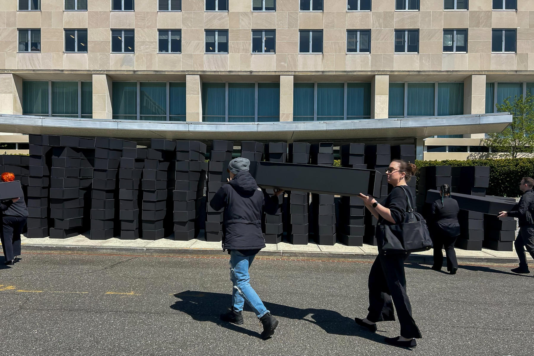 People carry mock coffins across a street toward a pile of hundreds of mock coffins in front of a building.