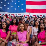 Several Black women in pink dresses sit together under an American flag.