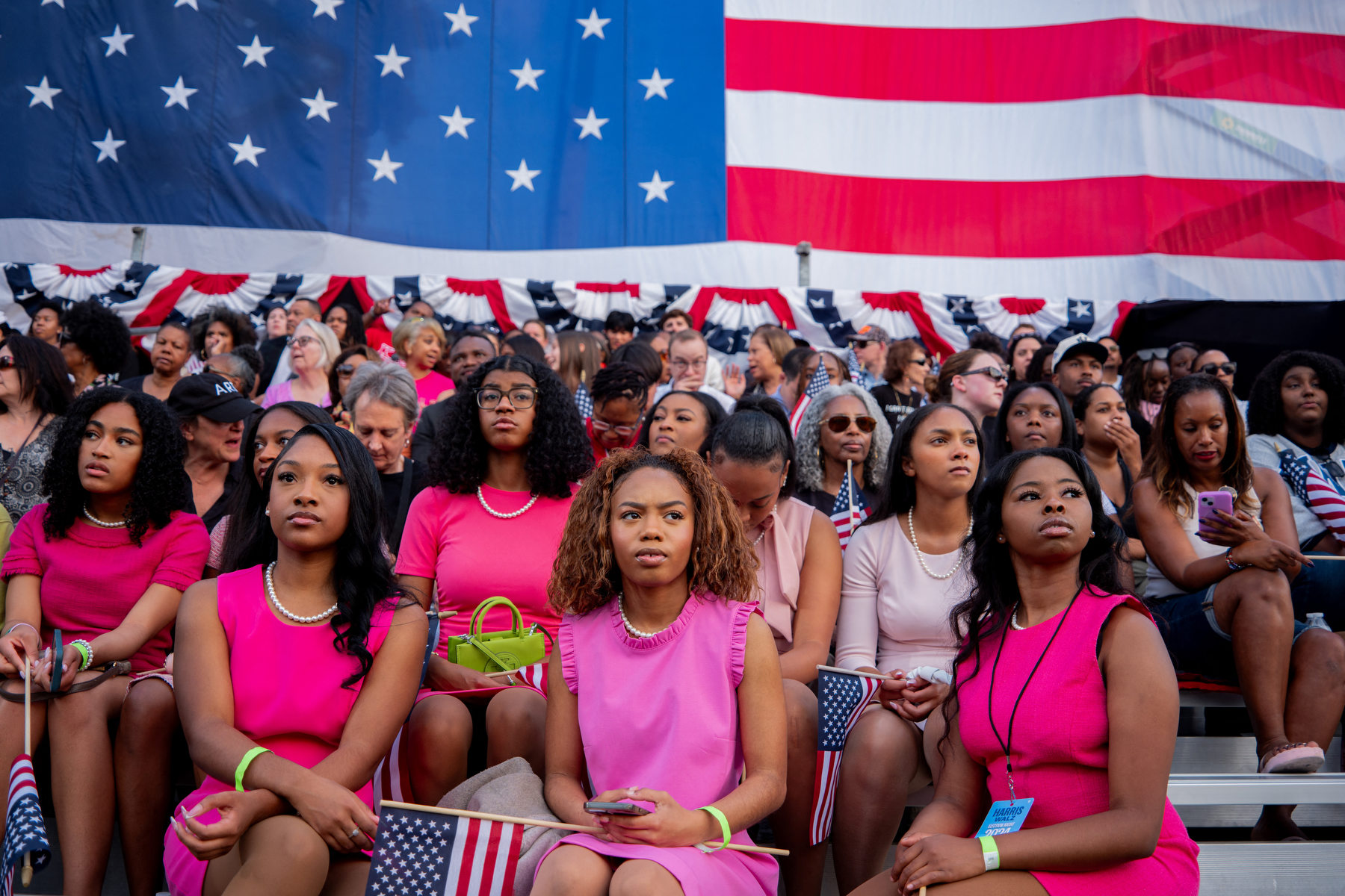 Several Black women in pink dresses sit together under an American flag.