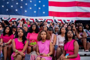 Several Black women in pink dresses sit together under an American flag.