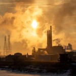 Industrial smokestacks emit thick smoke into the sky at sunset, casting a golden haze over the landscape.