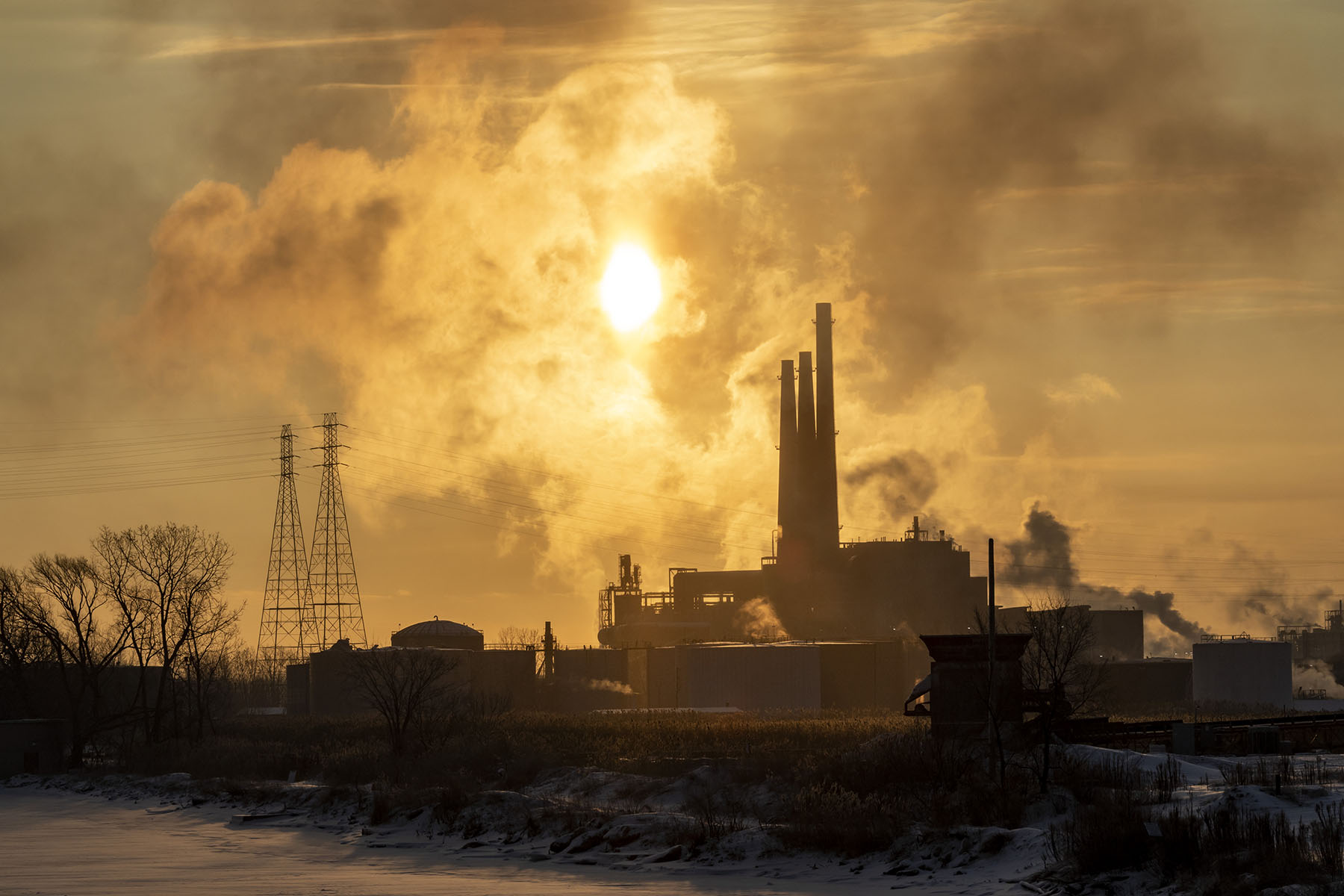 Industrial smokestacks emit thick smoke into the sky at sunset, casting a golden haze over the landscape.