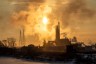 Industrial smokestacks emit thick smoke into the sky at sunset, casting a golden haze over the landscape.