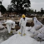 People wearing haz-mat suits and gear put burned materials in plastic bags.