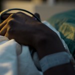 Close-up of a person's hand resting on a hospital bed, with a medical wristband and monitoring clip attached to their finger.