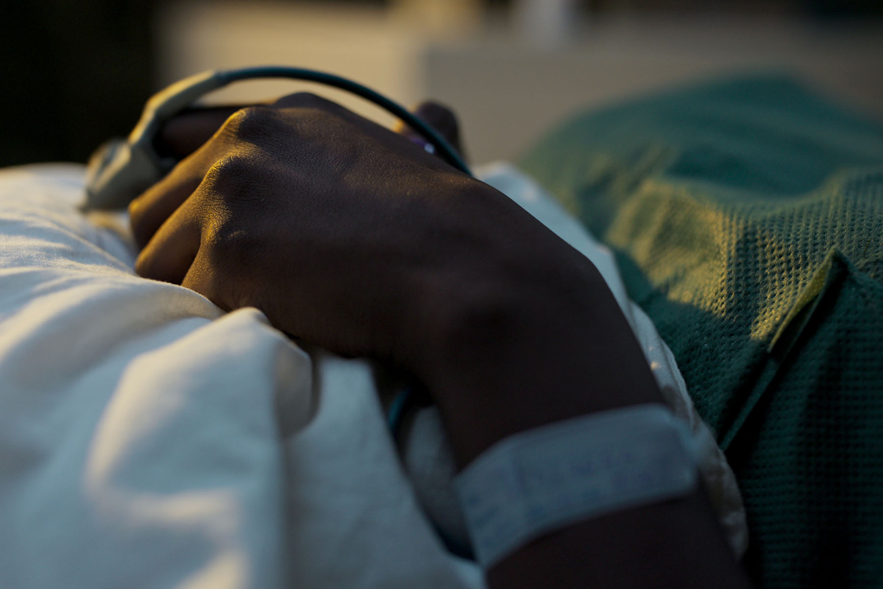 Close-up of a person's hand resting on a hospital bed, with a medical wristband and monitoring clip attached to their finger.