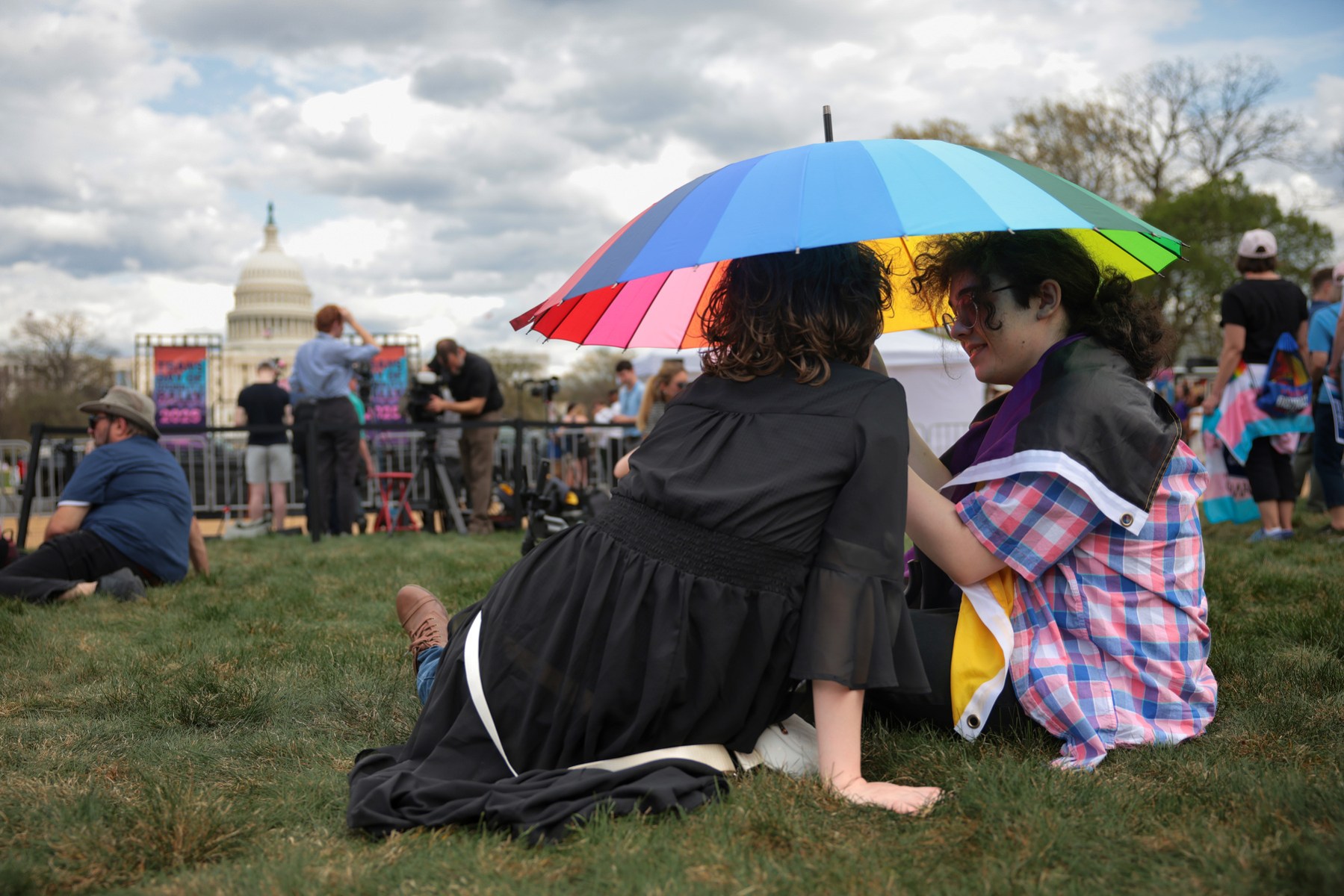 Two people sit on the grass under a colorful umbrella.