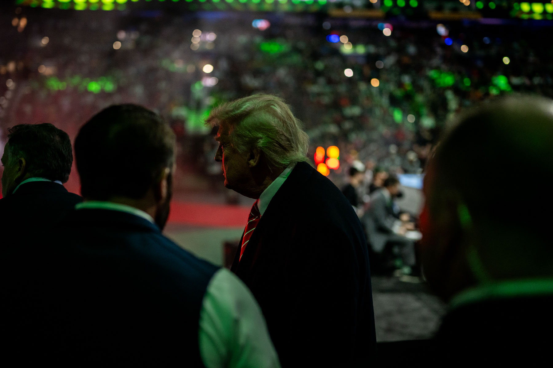 Donald Trump stands in shadow, his face partially illuminated by green lighting, looking out over a packed UFC arena filled with fans and flashing lights.