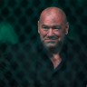 UFC President Dana White looks through the chain-link of an octagon cage, smiling slightly.