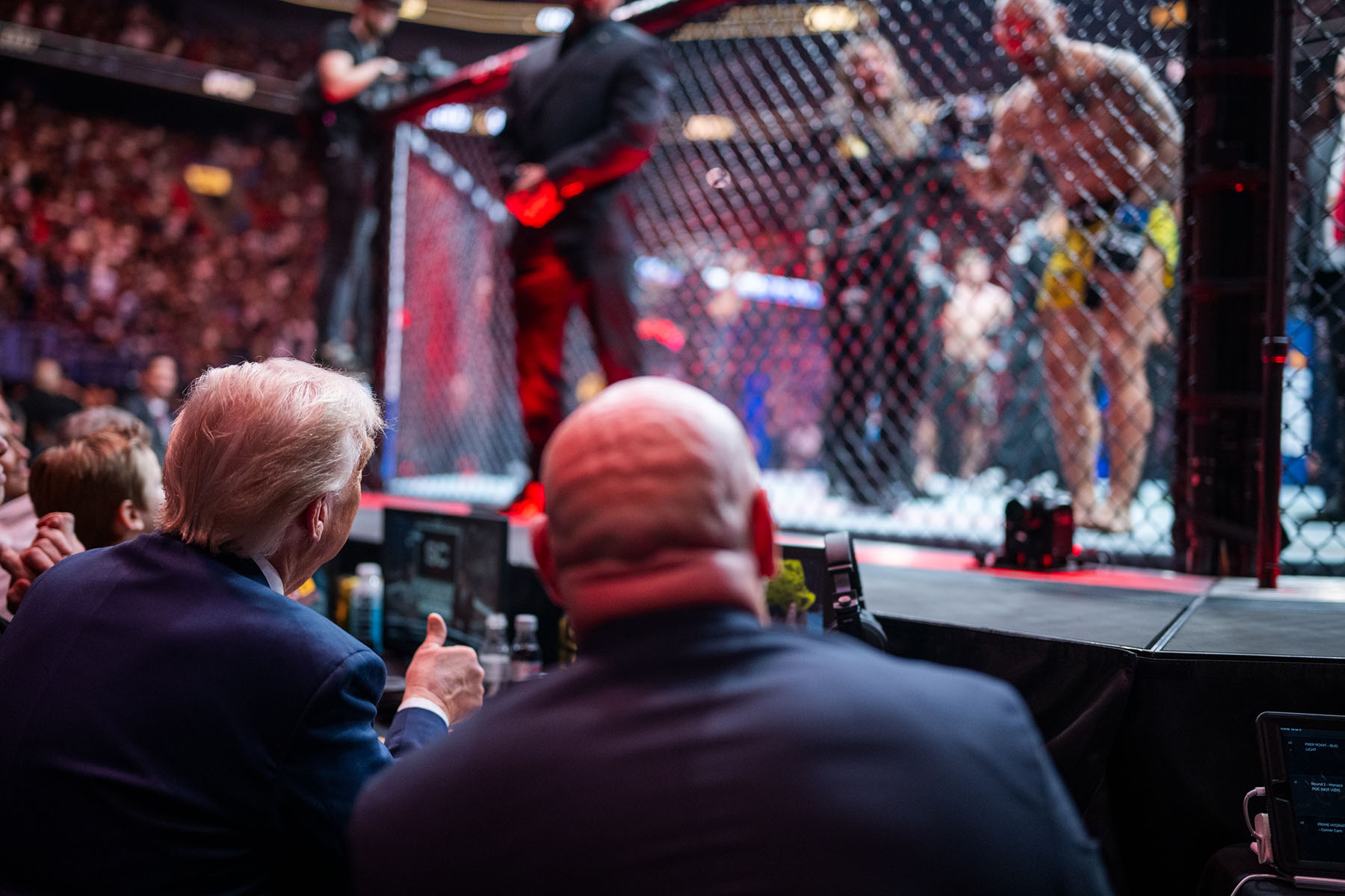 Donald Trump and Dana White sit cageside at a UFC match, watching fighters in the octagon, with Trump giving a thumbs-up and the cage brightly lit in red.