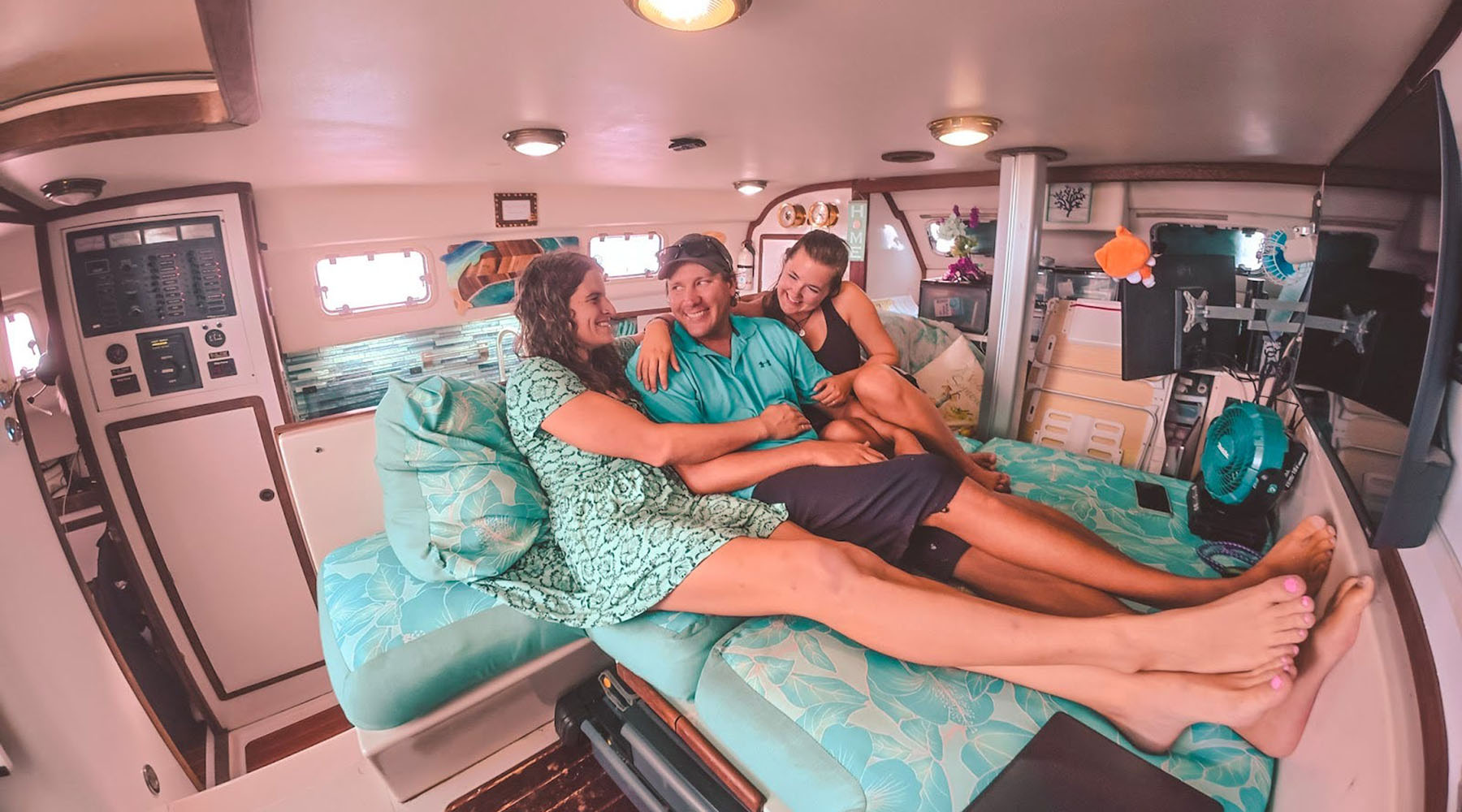 Three people lounge and laugh together on a teal-cushioned bed inside a boat cabin.