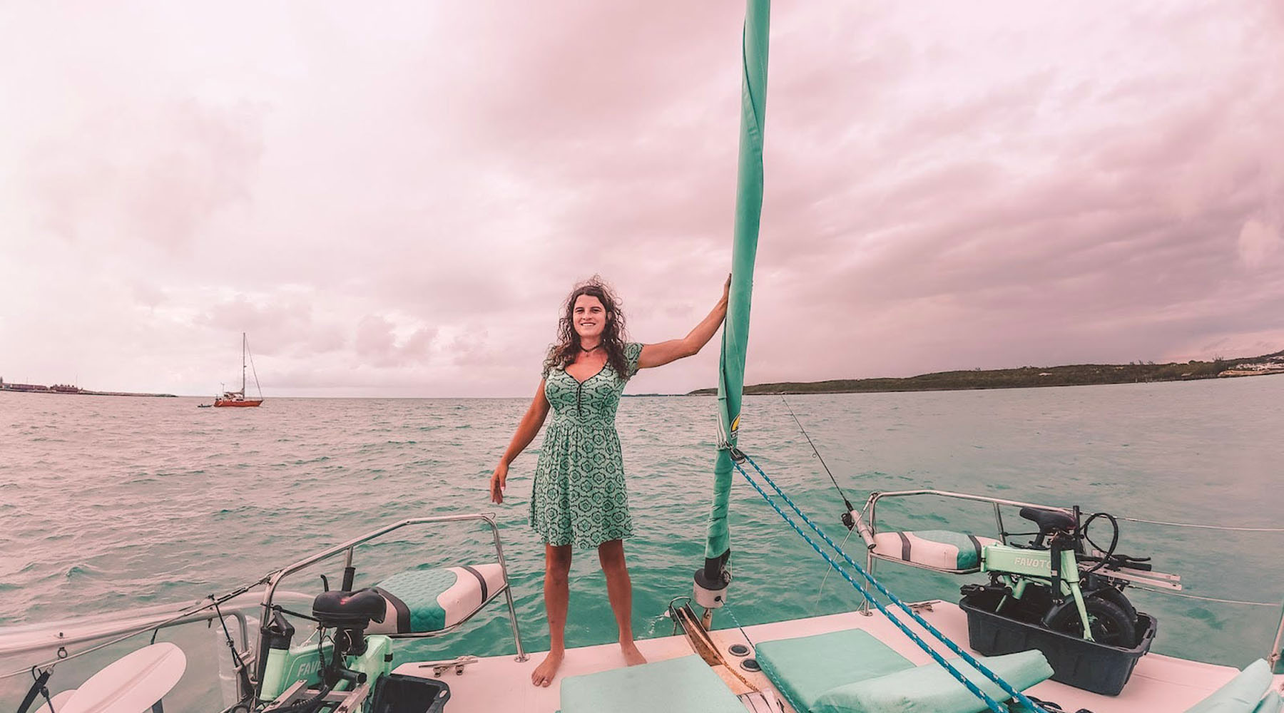 Kelsey Granger stands barefoot on the deck of a sailboat, holding onto a green sail with one hand and smiling toward the horizon.