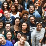A large, diverse group of young people smiles up at the camera in a brightly lit indoor setting.