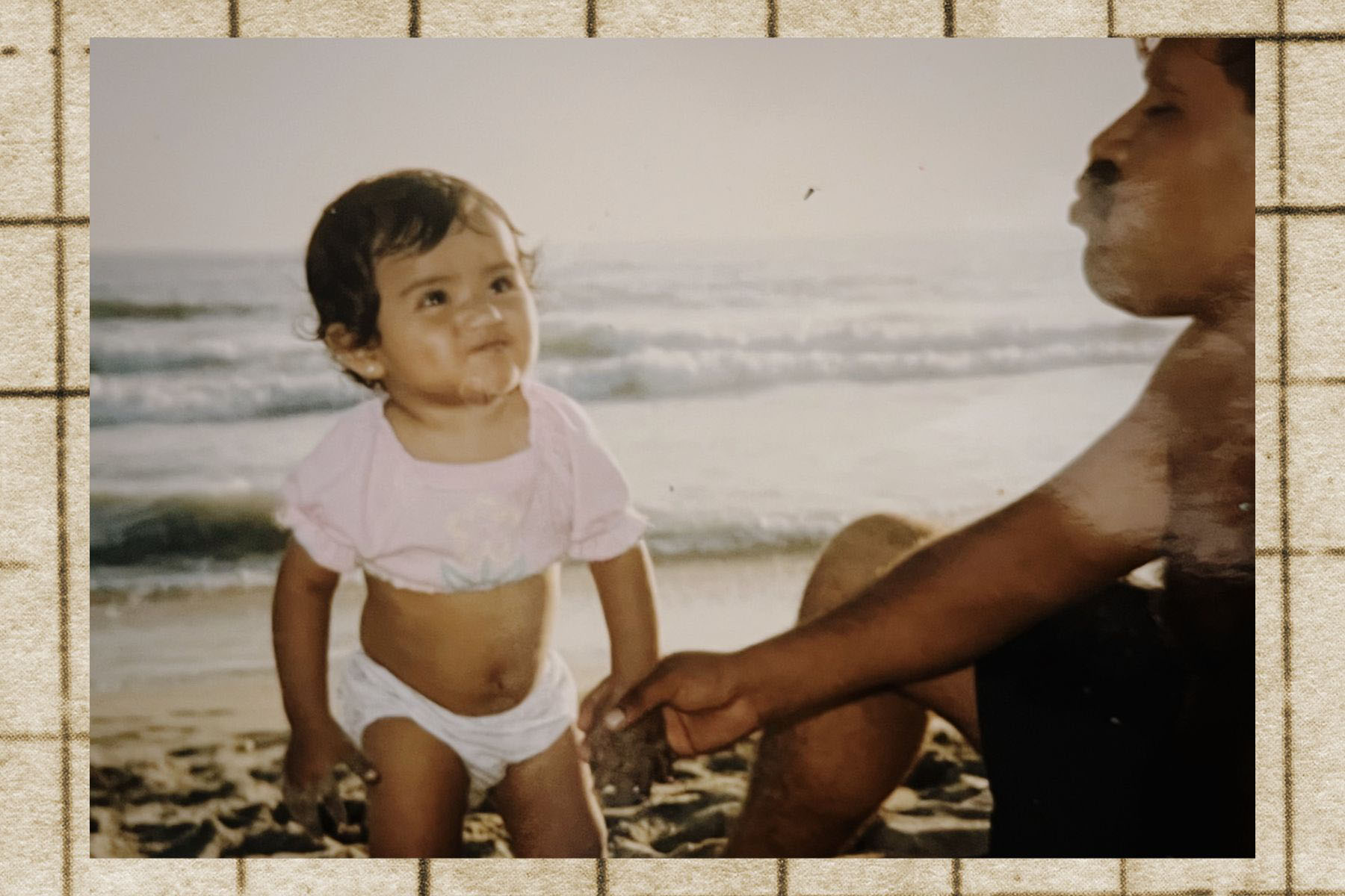 A toddler-aged Reyes stands in the sand at the beach, smiling toward her father, who is partially visible and holding her hand.